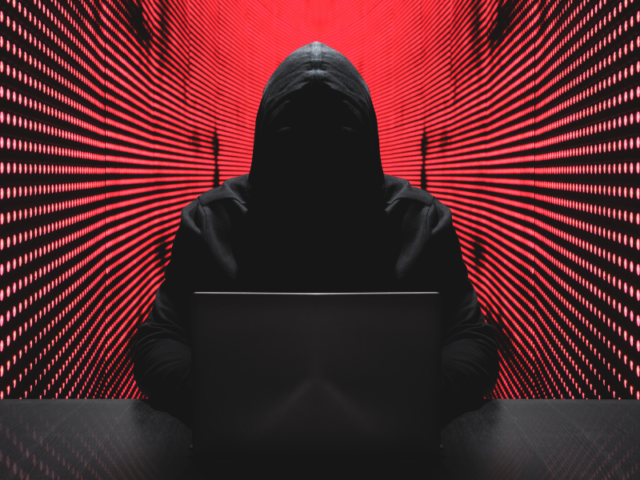 Image of a man in a hood behind a computer - his face is shrouded in darkness.