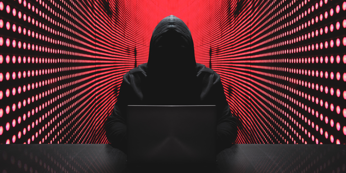 Image of a man in a hood behind a computer - his face is shrouded in darkness.