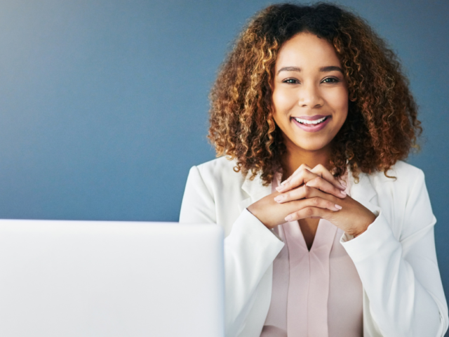 How to Start Doing Contract Work Blog Graphic - photo of a woman in a white blazer. She's sitting in front of a blue wall with her hands folded. She's smiling and sitting in front of a white laptop.